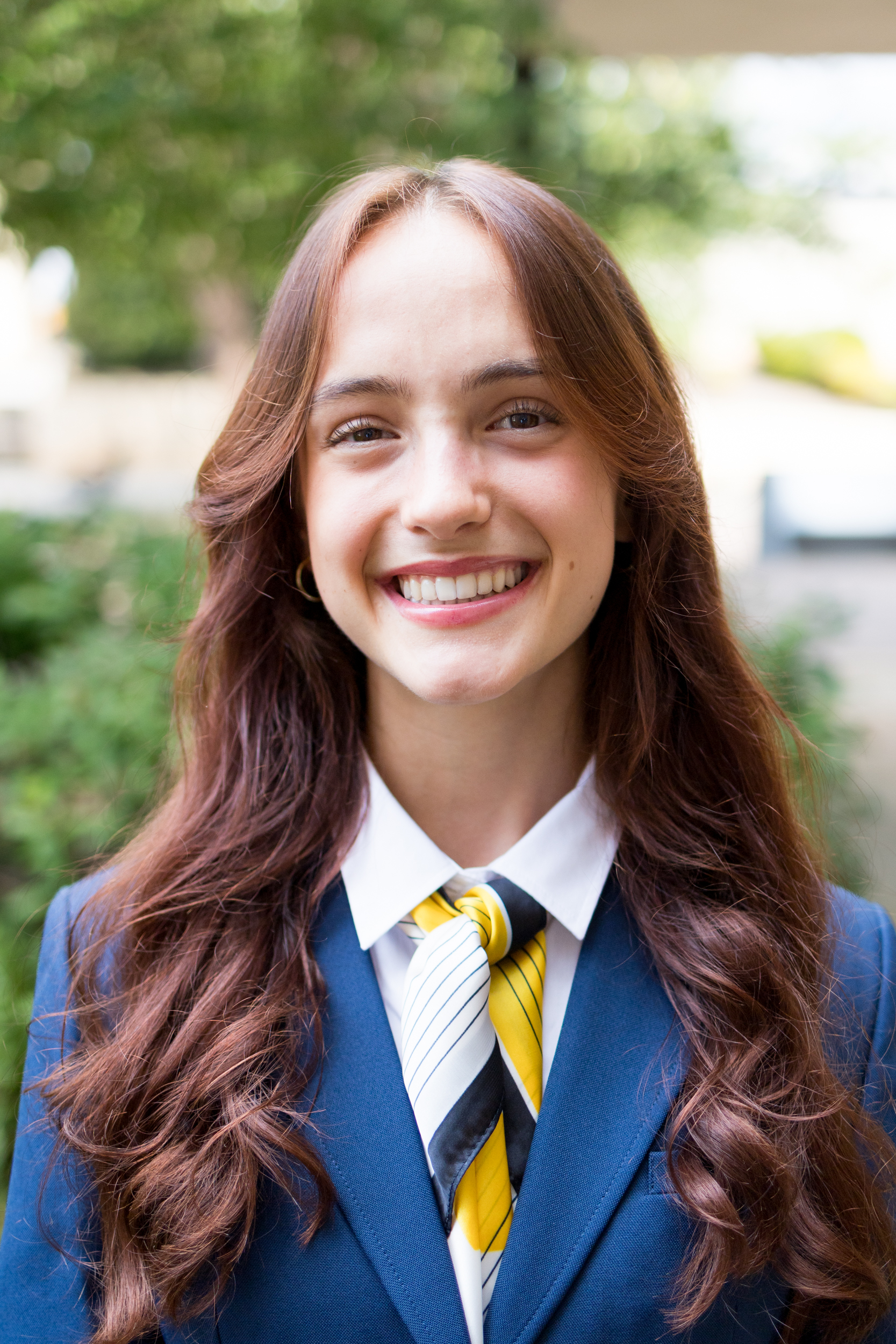 Destiny McPherson, wearing big black cat-eye glasses, has her two-tone brown hair pulled back with a black headband and a small braid pulled back on each side. She is dressed in a navy blazer, a white shirt, a navy and gold scarf, and hanging wooden earrings, completed by a smile.