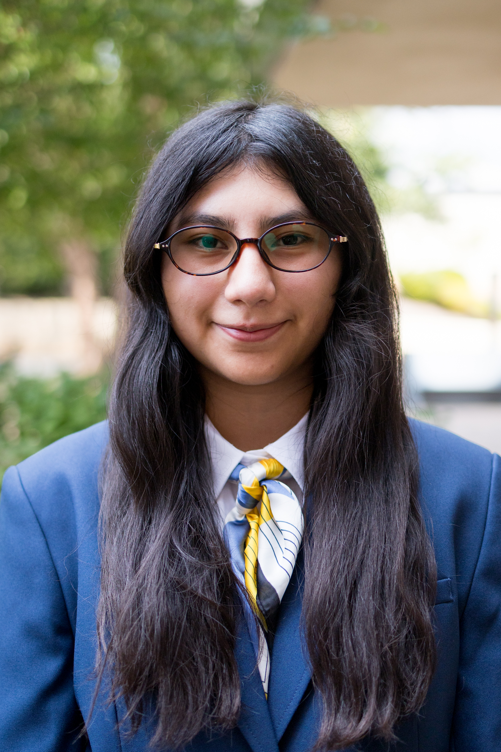 Kaitlyn Vanderburg wearing red-framed glasses and has shoulder-length hair. Her outfit includes a white collared shirt with a dark blue blazer. She also wears a scarf with a pattern of navy blue, yellow stripes, and blue polka dots. The background consists of a brick wall with a slightly blurred appearance, emphasizing the focus on the subject.<br />